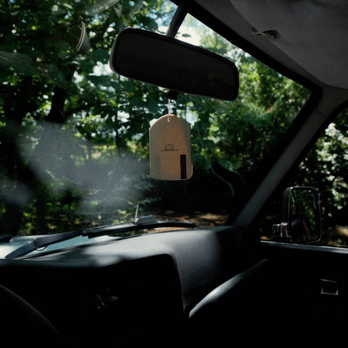 A car cologne card hanging from a car's rearview mirror, with a forest visible through the windshield.